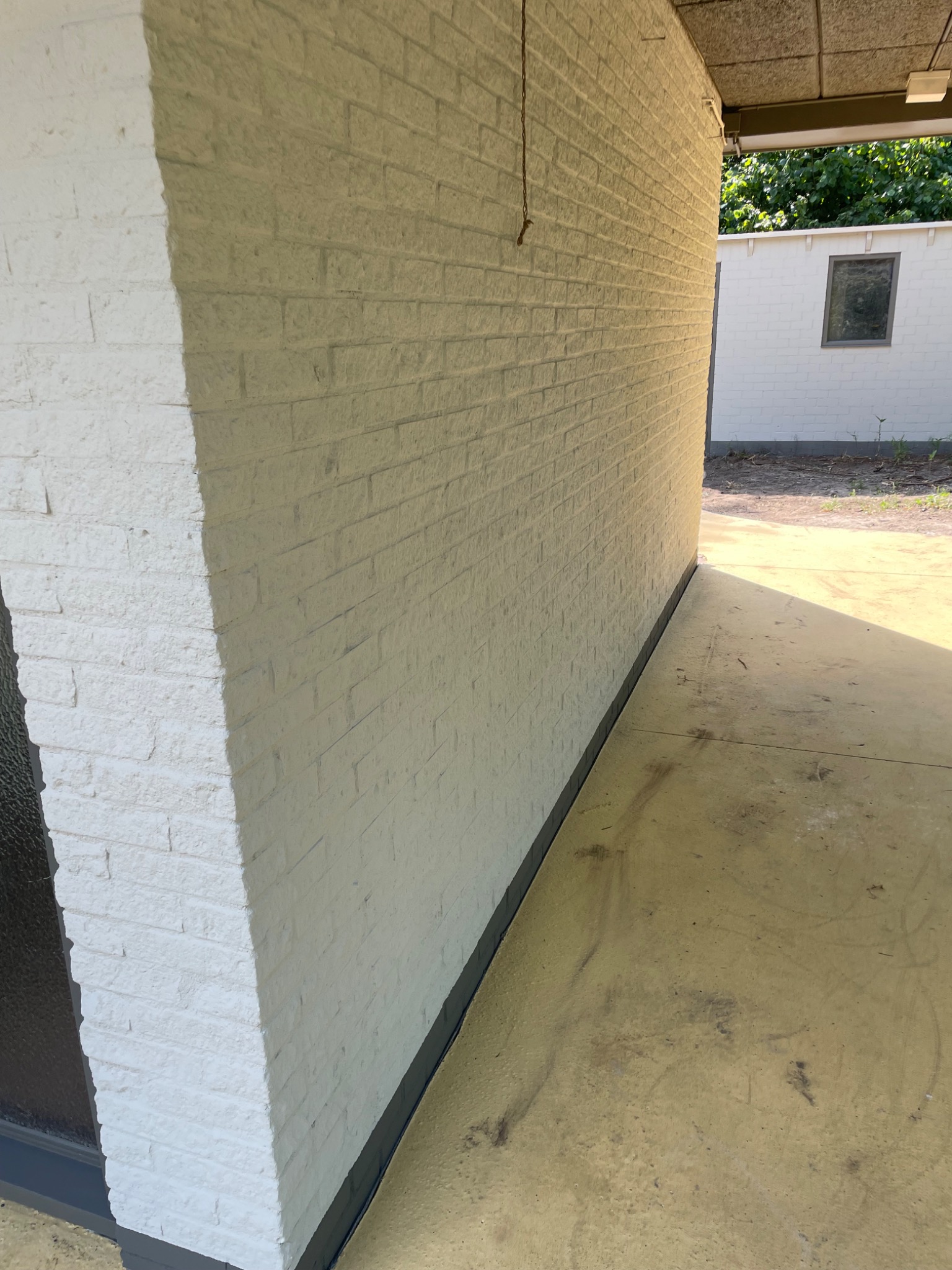 Biała, pomalowana ceglana ściana zewnętrzna budynku, widoczna z boku, z fragmentem okna po lewej stronie i betonową podłogą, z małym budynkiem w tle.