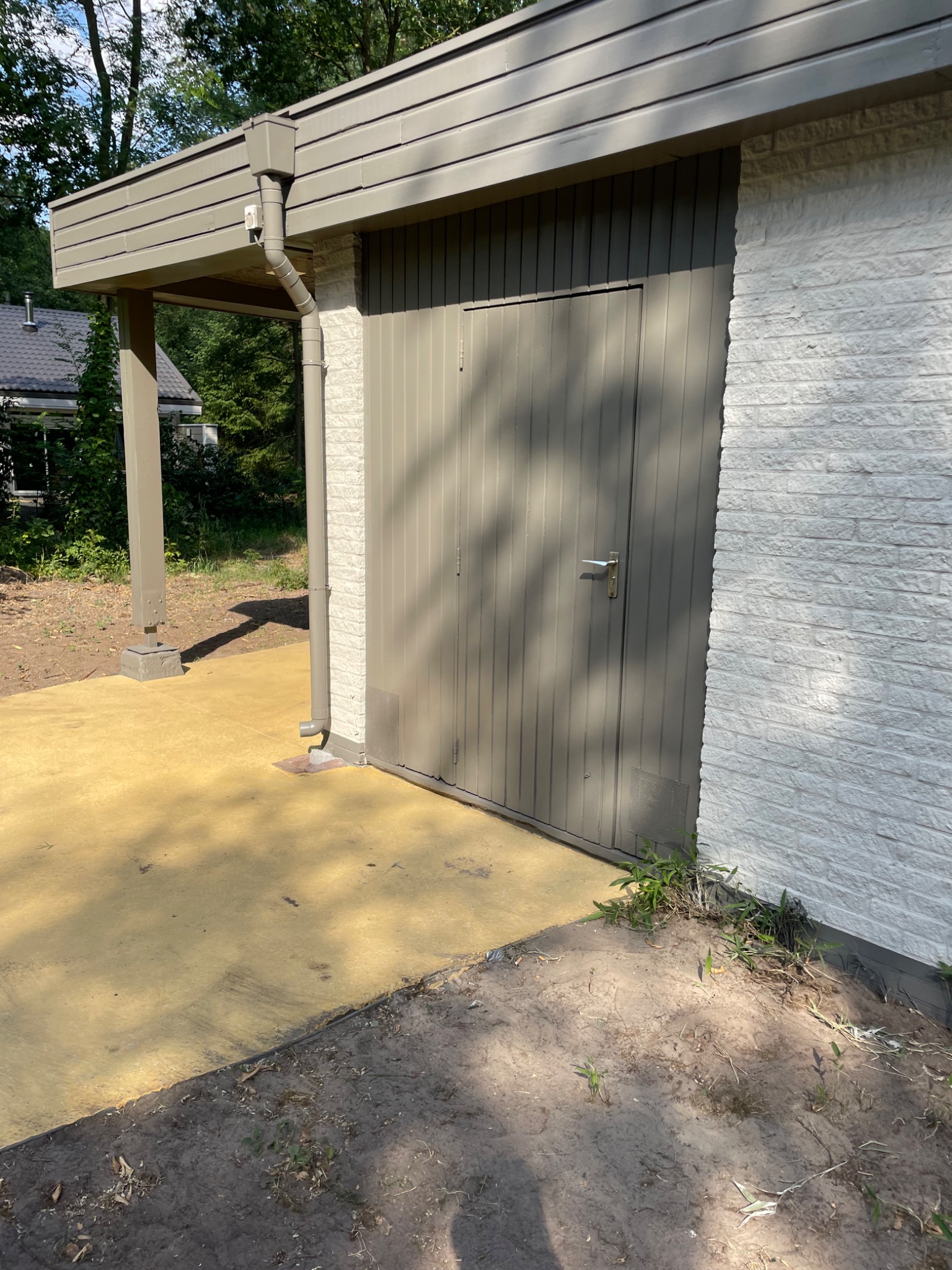 Pomieszczenie gospodarcze z pomalowaną elewacją z cegły, szarą bramą i świeżo wylaną, żółtą posadzką z betonu.