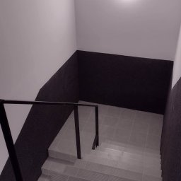 Krkrkr - Klatka schodowa wykończona płytkami ceramicznymi w odcieniach szarości, z czarną balustradą i częściowo pomalowanymi ścianami na biało i szaro, oświetlona okrągłą lampą sufitową.