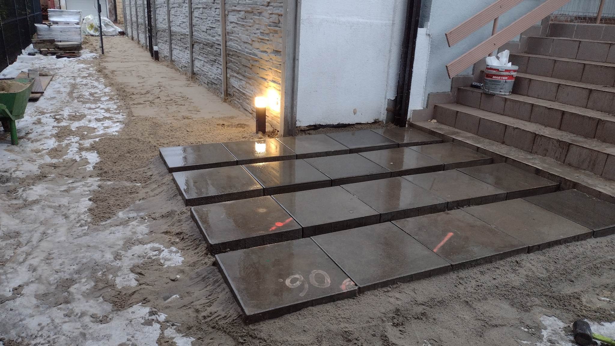 Układanie stopni z ciemnych, kwadratowych płyt chodnikowych prowadzących do wejścia, widoczny piasek, lampa ogrodowa i wiadro z farbą na schodach.