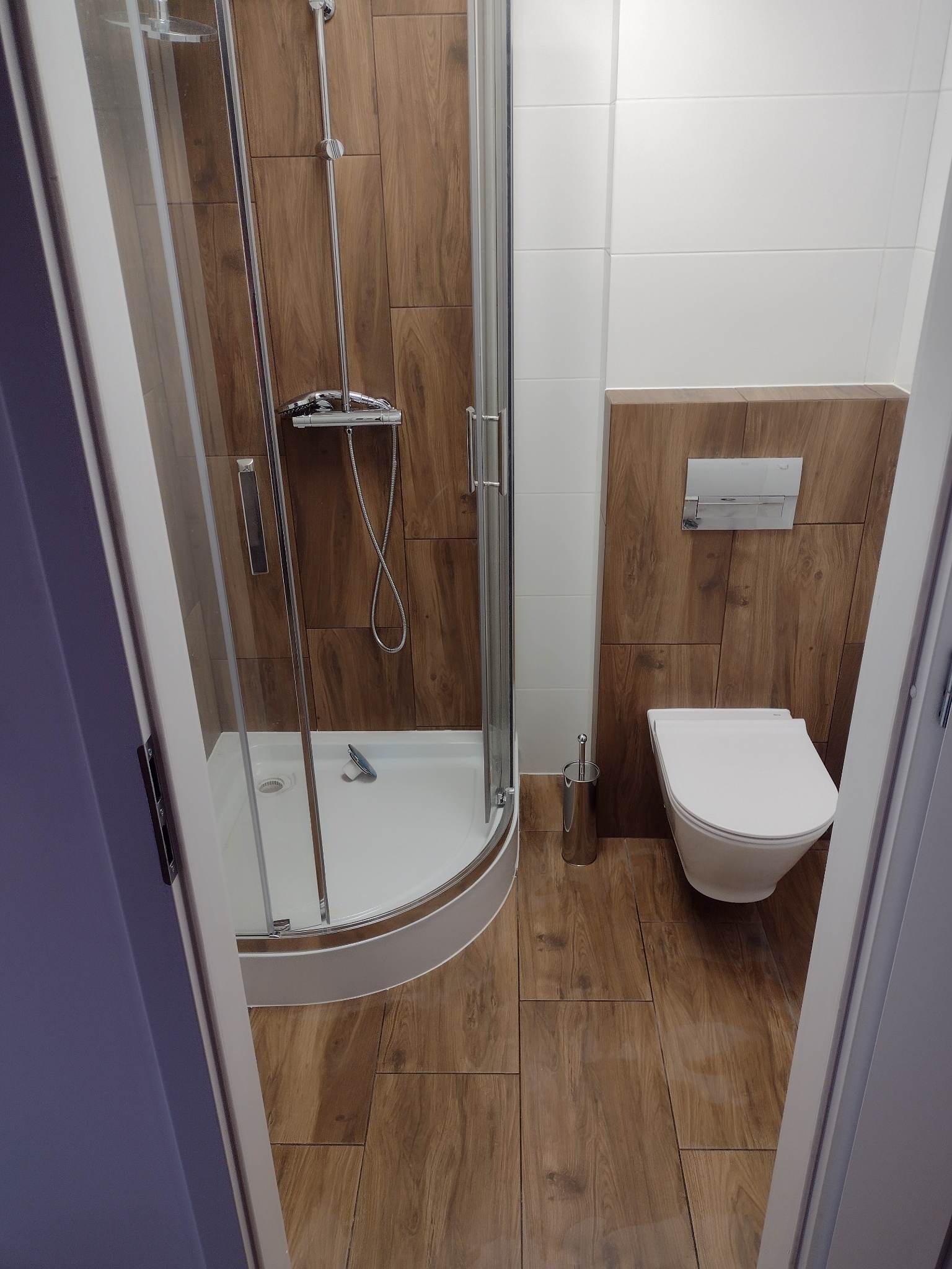 Wykończona łazienka z kabiną prysznicową z zaokrąglonym brodzikiem, podwieszaną toaletą i drewnopodobnymi płytkami na podłodze i części ścian, widok zza uchylonych drzwi.