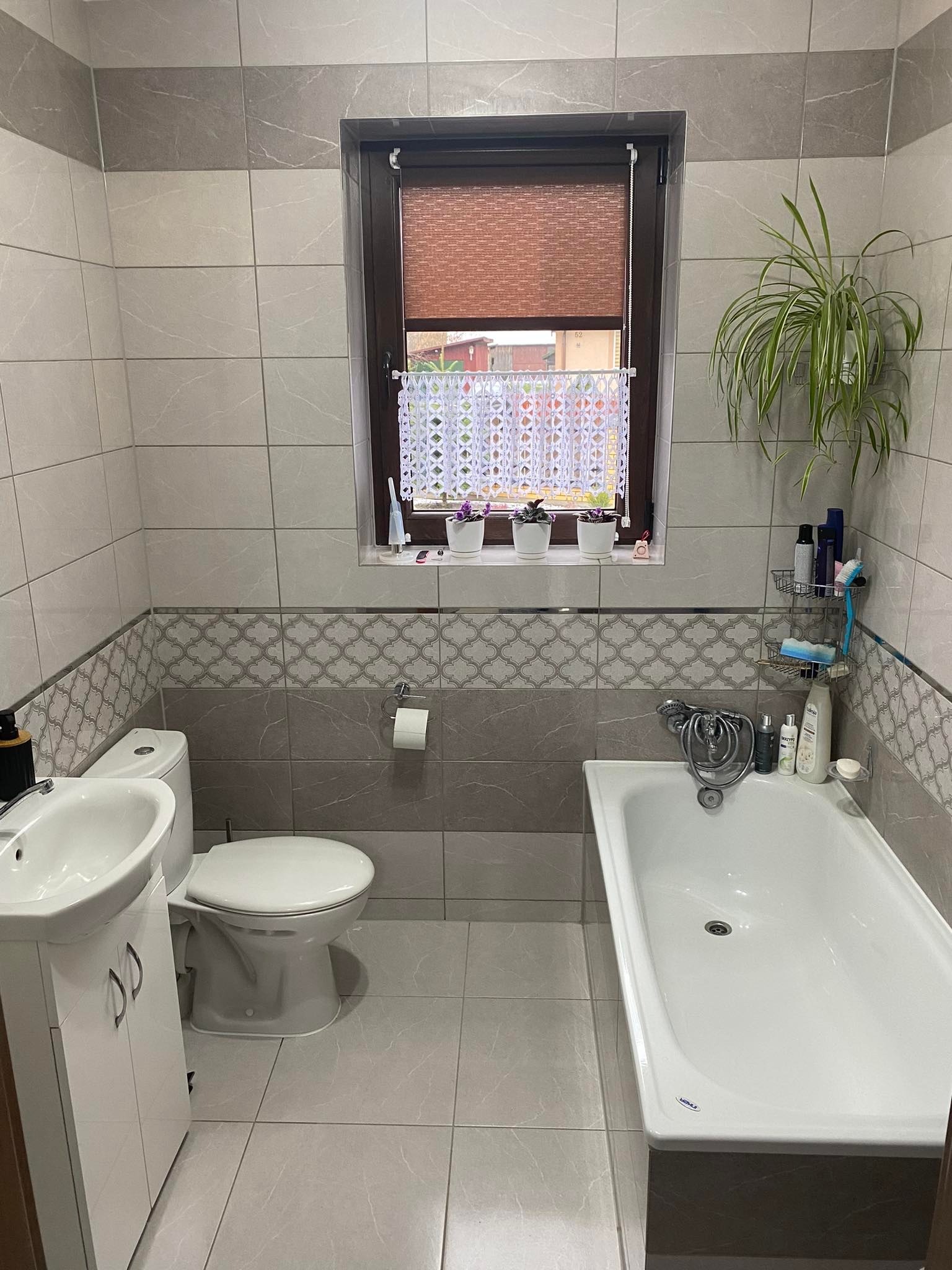 Wyremontowana łazienka z szaro-białymi płytkami, białą armaturą, oknem z roletą i roślinami doniczkowymi, widok z przodu.