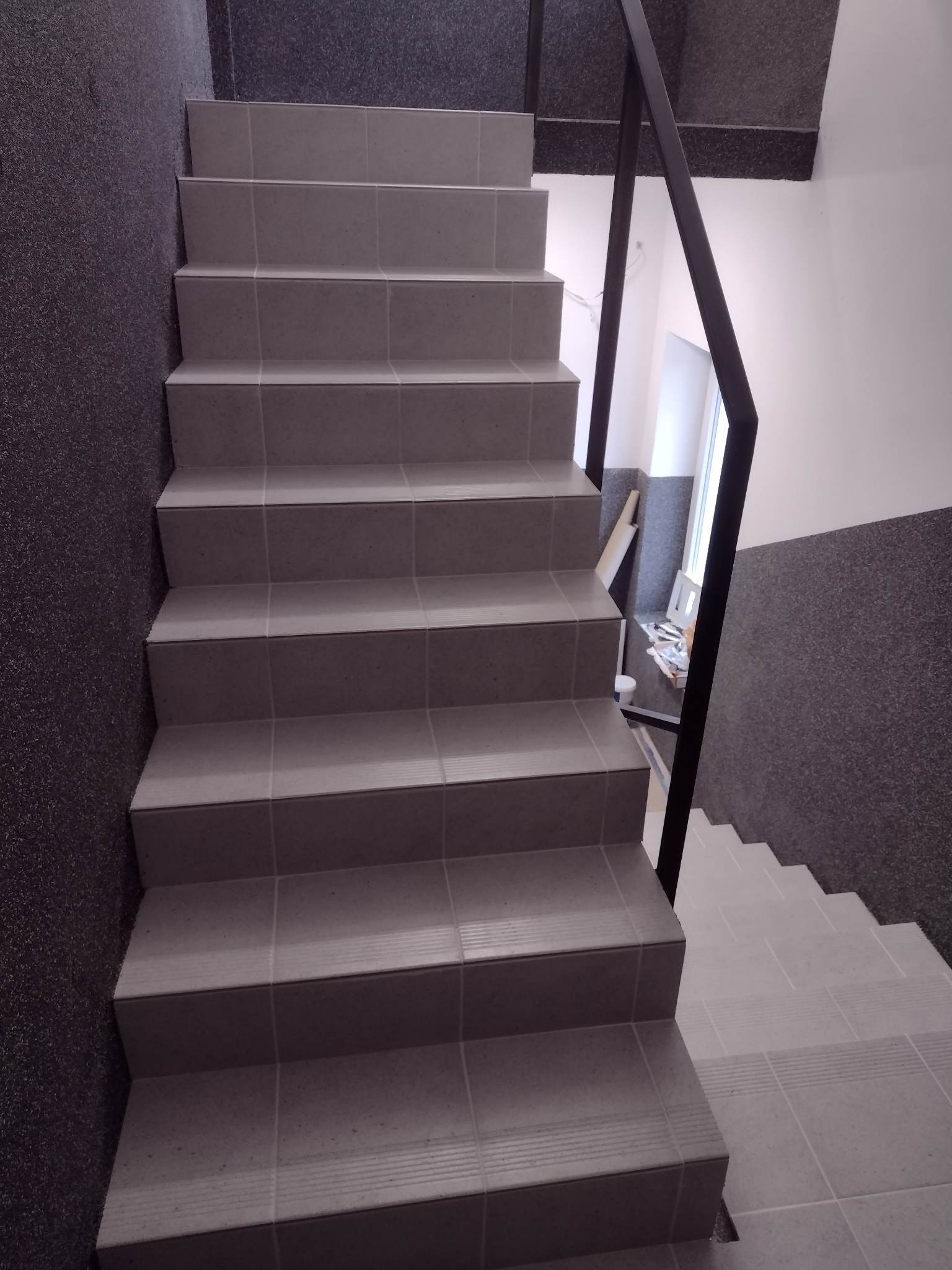 Klatka schodowa z jasnoszarymi, kwadratowymi płytkami na stopniach, widoczne czarne metalowe poręcze, ściany w dwóch odcieniach szarości, dolne schody wykończone paskami antypoślizgowymi.