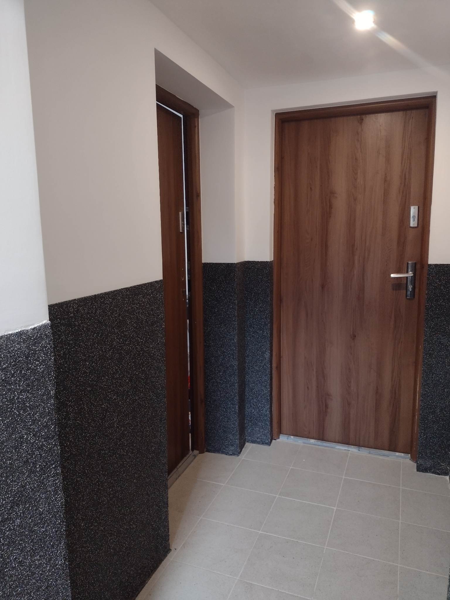 Korytarz po remoncie: brązowe drzwi wejściowe z metalową klamką i wizjerem, obok drugie drewniane drzwi, ściany pomalowane w górnej części na jasny beż, dolna część pokryta czarnym, chropowatym...