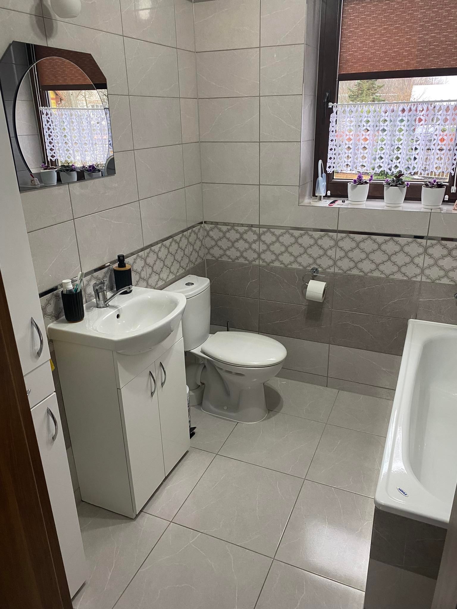 Jasna łazienka z szarymi płytkami imitującymi marmur, białymi meblami łazienkowymi, lustrem z czarną ramą i dekoracjami okiennymi w postaci firanki i kwiatów w doniczkach.