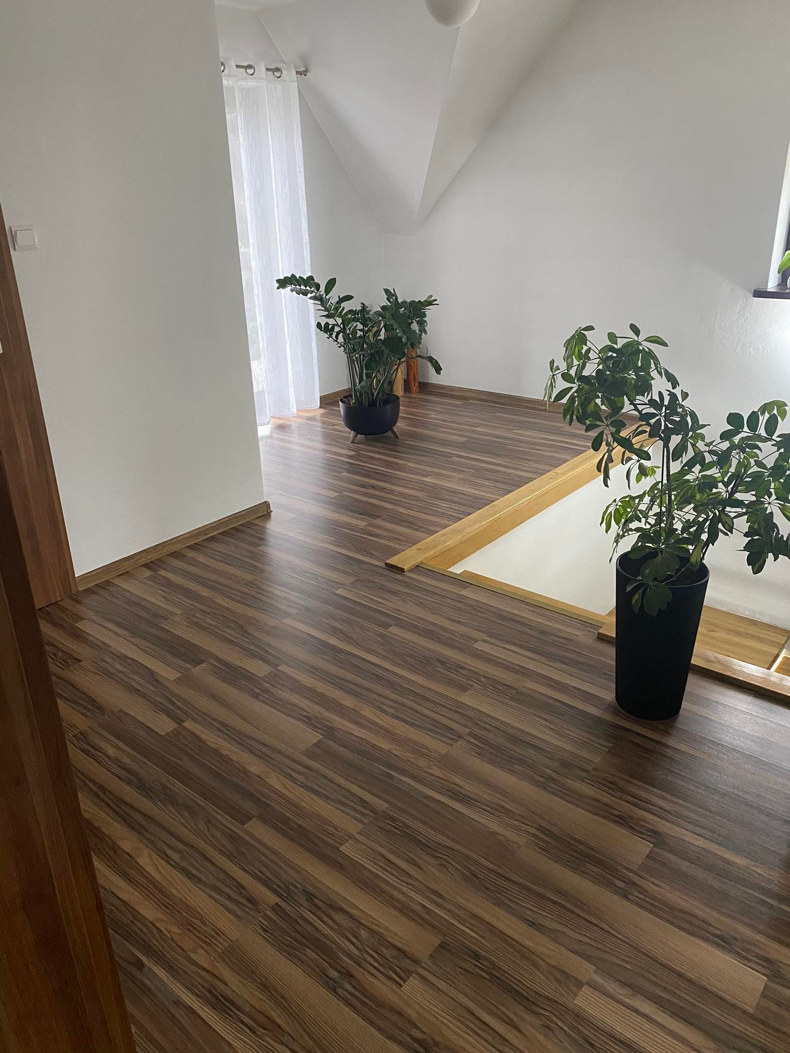 Pokój na poddaszu z brązową podłogą imitującą drewno, świeżo pomalowane białe ściany, drewniane listwy przypodłogowe i dwie duże rośliny doniczkowe.