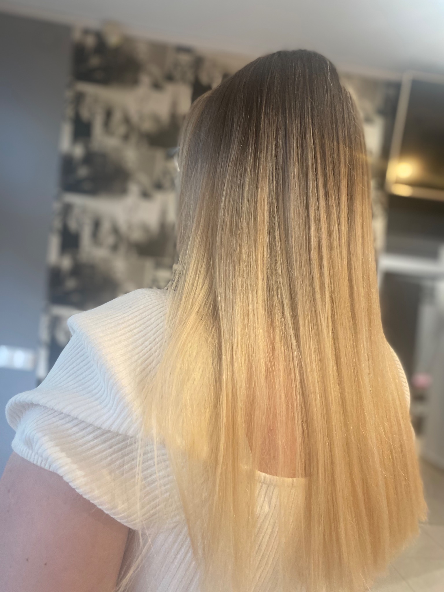 Długie, proste włosy blond z ciemniejszym odrostem, widok z tyłu, na tle ściany z fototapetą.