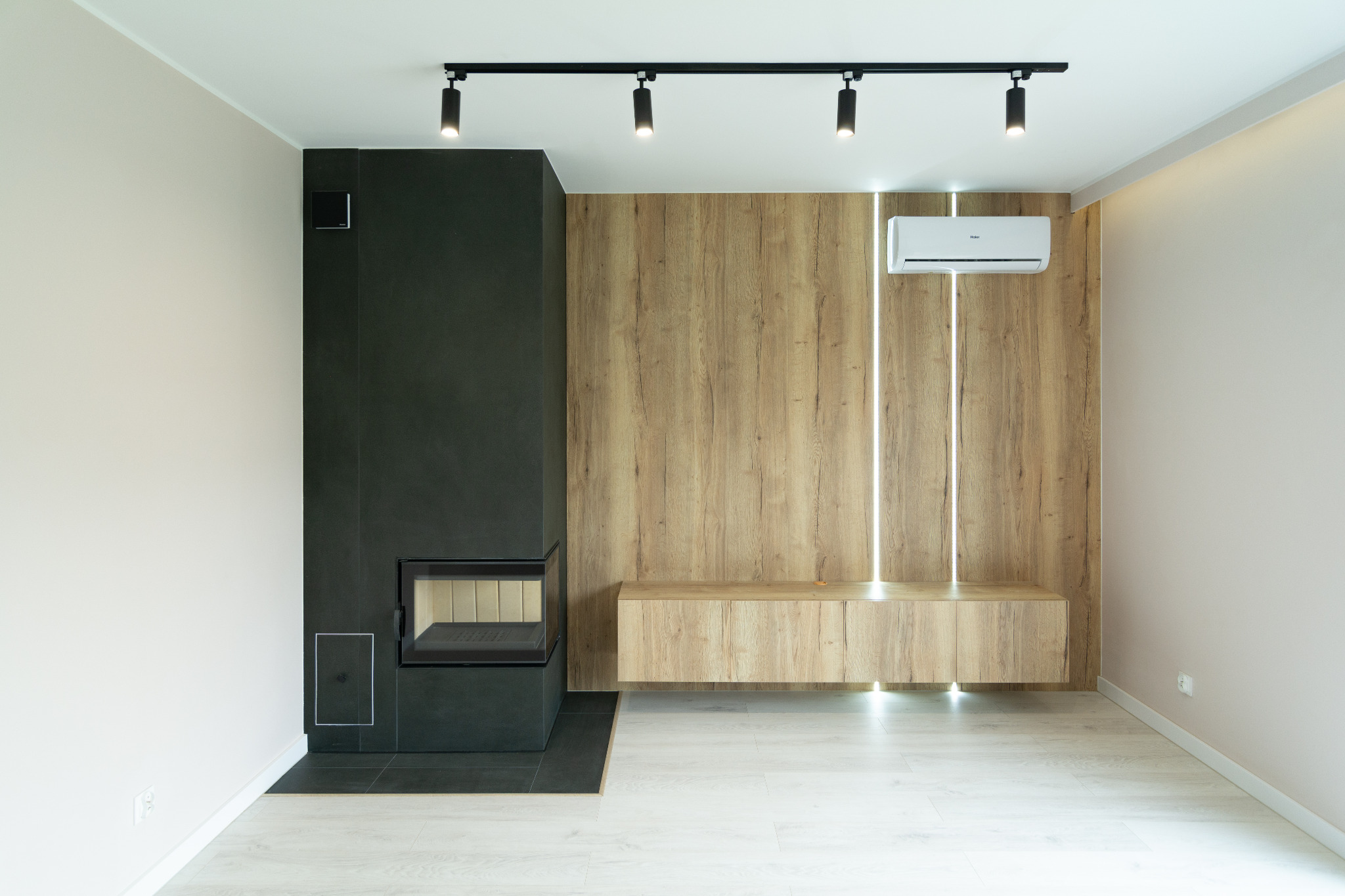 Nowoczesny salon z czarnym kominkiem, drewnianą ścianą z podświetleniem LED i klimatyzatorem, oświetlenie punktowe na suficie, jasna podłoga.