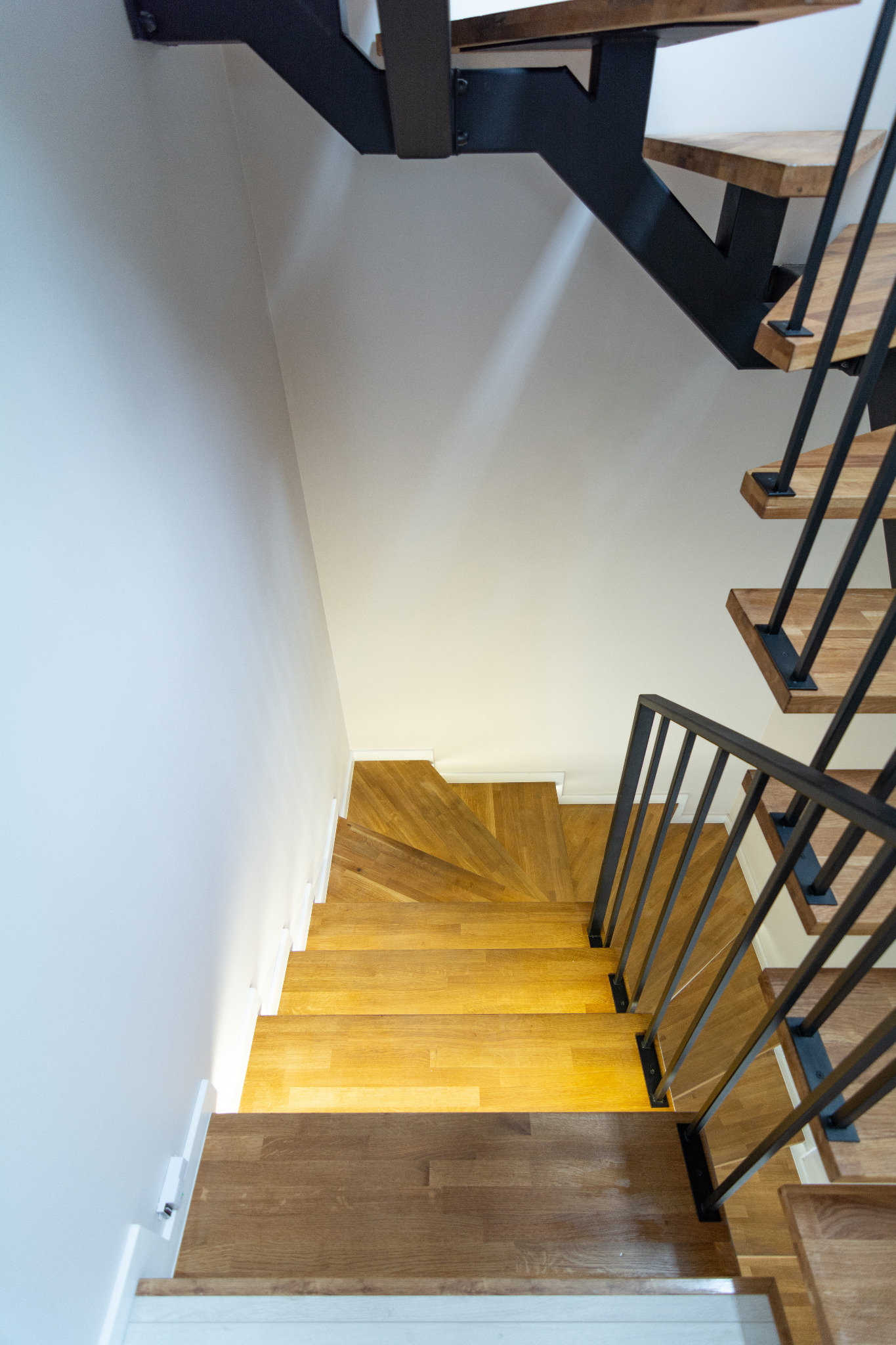 Widok z góry na drewniane schody z czarną metalową konstrukcją i minimalistyczną białą ścianą, ukazujący nowoczesny design i precyzję wykonania.