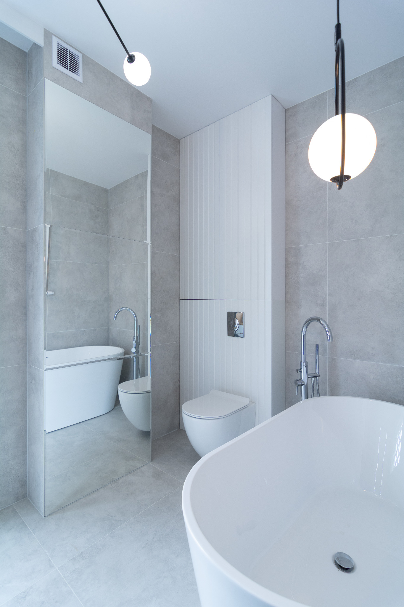Minimalistyczna łazienka w szarościach z wolnostojącą wanną i toaletą typu Geberit, odbicie w lustrze ukazuje dodatkową wannę, oświetlona wiszącą lampą kulą.