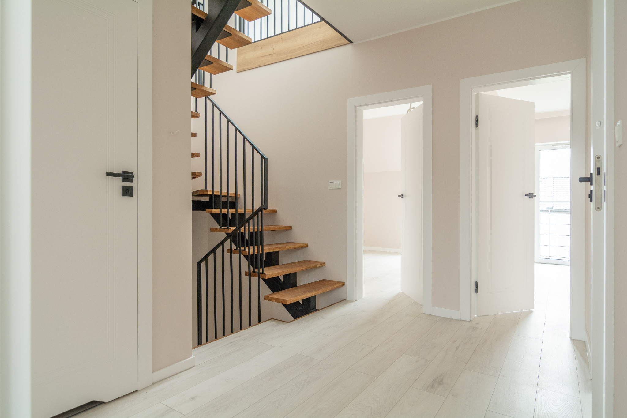 Nowoczesne wnętrze z drewnianymi schodami z czarną balustradą, białymi drzwiami z czarnymi klamkami i jasną podłogą, widok na otwarte drzwi do kolejnych pomieszczeń.