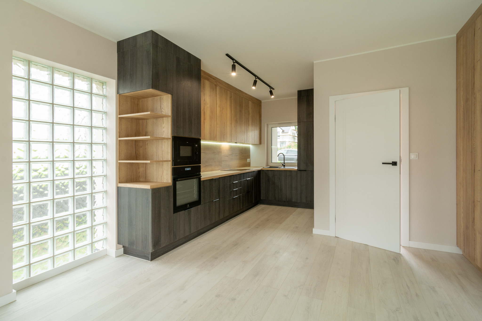 Nowoczesna kuchnia z drewnianymi szafkami w odcieniach szarości i dębu, oświetleniem punktowym i oknem z luksferami, wykończona jasnymi panelami podłogowymi.