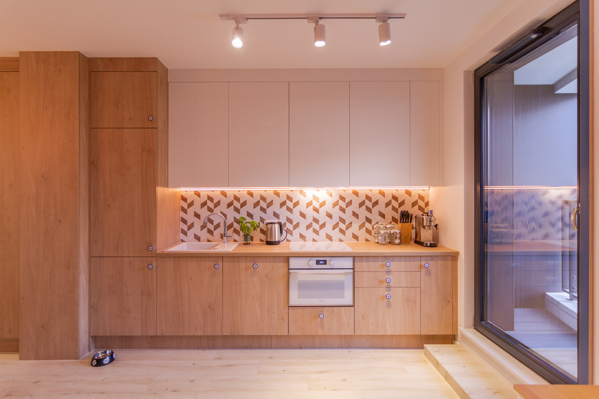 Jasna kuchnia z drewnianymi frontami, mozaiką w jodełkę nad blatem i oświetleniem punktowym, widok na ogród przez duże okno.