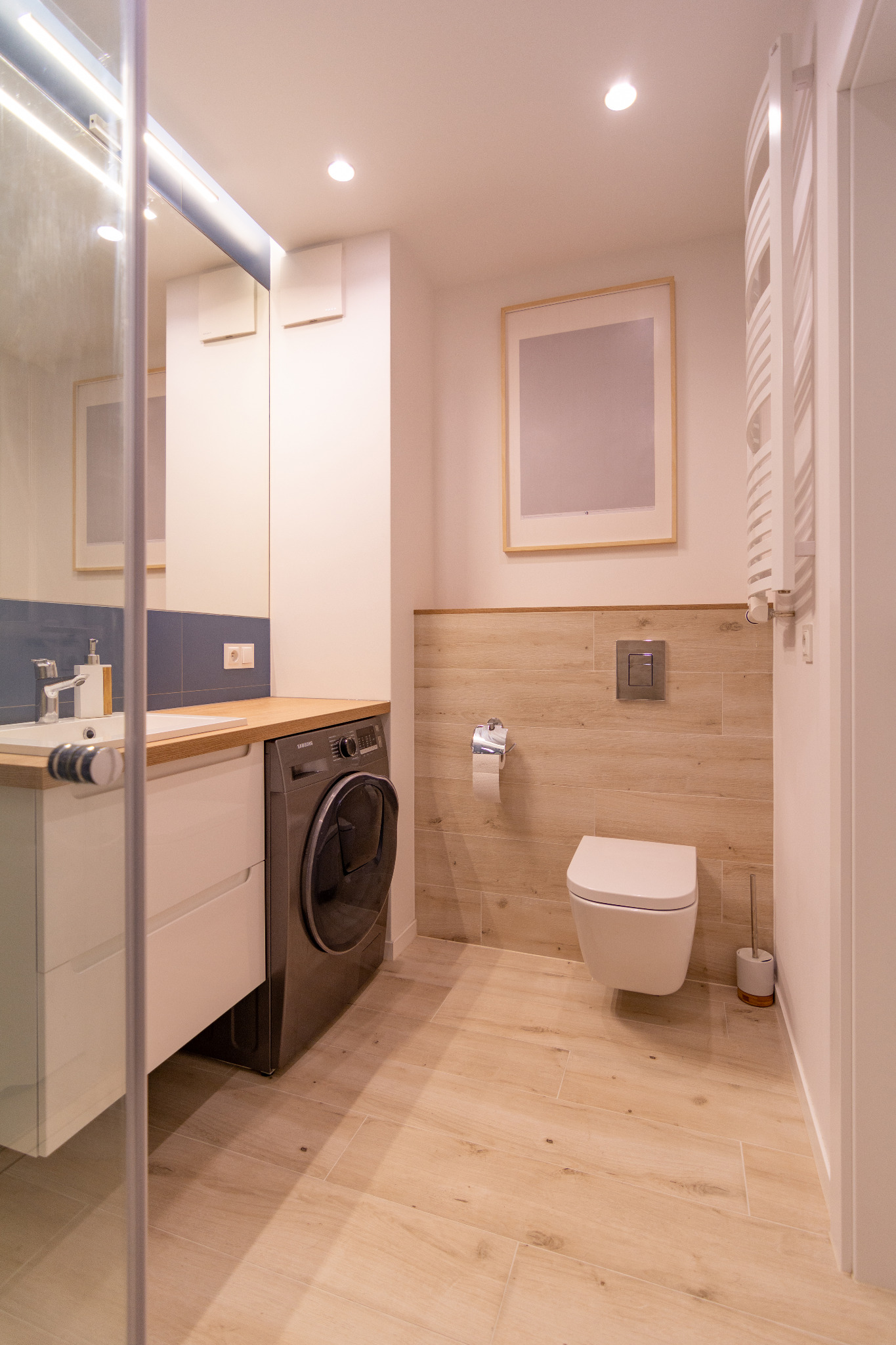 Jasna łazienka z drewnopodobnymi płytkami na podłodze i ścianie, biała umywalka z szafką i drewnianym blatem, pralka pod blatem, toaleta podwieszana i grzejnik drabinkowy.
