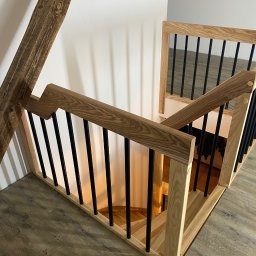 Vvwood szczecin - Spiralne schody z jasnego drewna z czarnymi, metalowymi prętami balustrady, widoczne z góry, z fragmentem belki stropowej w lewym górnym rogu.