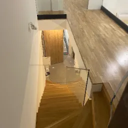 Wnętrze domu z drewnianymi schodami i szklaną balustradą. Widok z góry na dół, ukazujący drewniane stopnie i podłogę, oraz fragment drzwi wejściowych.