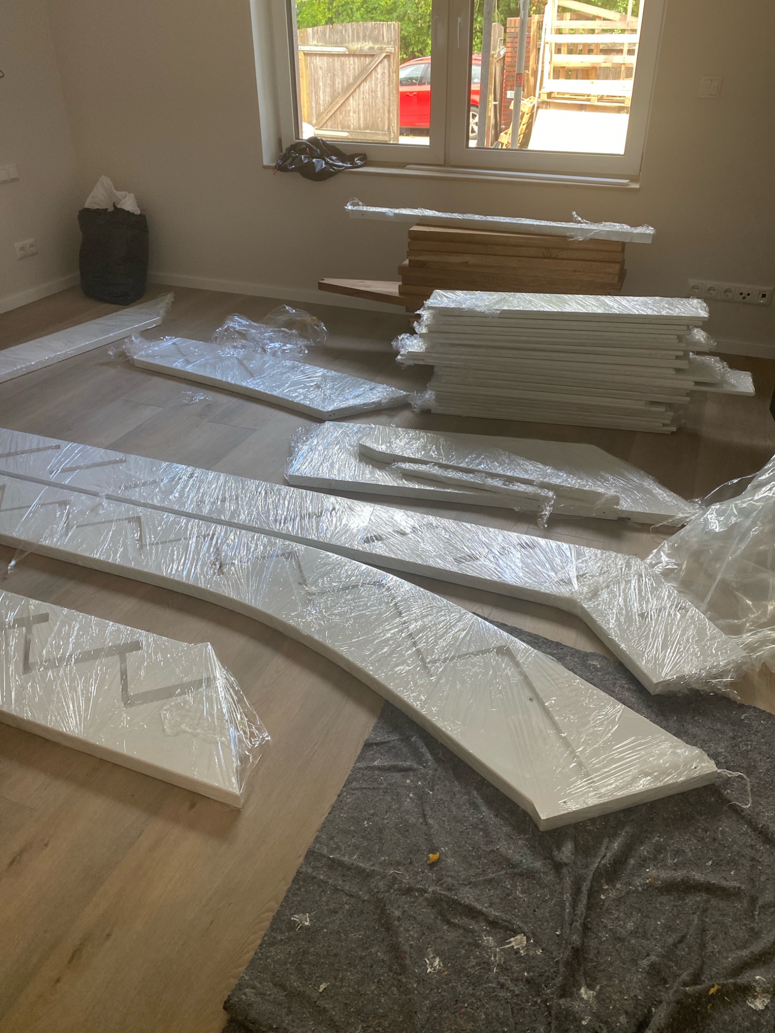 Białe elementy schodów, owinięte folią, leżą na podłodze w pomieszczeniu. Widoczne deski, stopnie i fragmenty balustrady. Częściowo widoczne okno i śmieci.