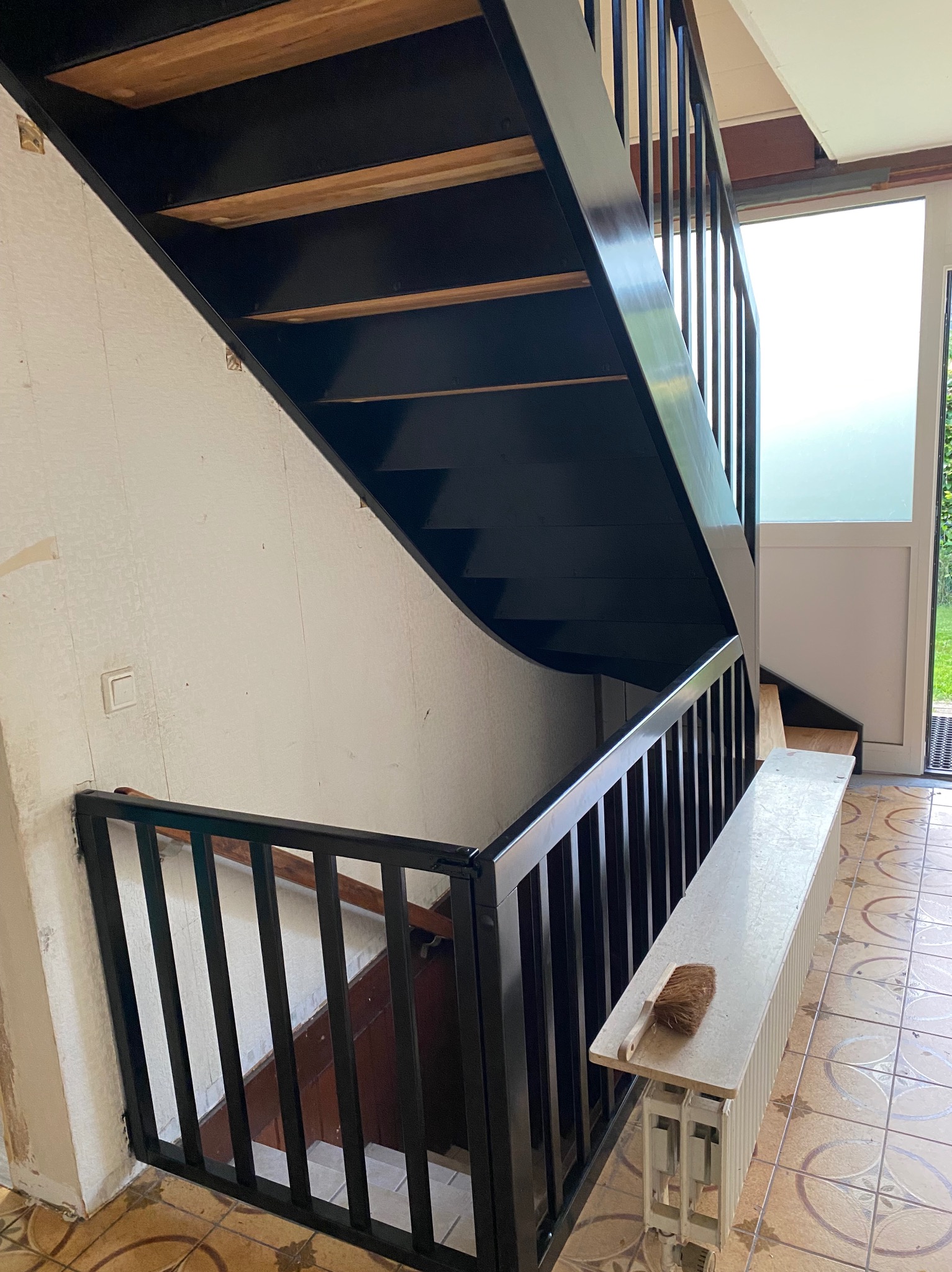 Czarne schody drewniane z balustradą, widok z dołu, z drewnianymi stopniami i białą ścianą w tle. Na grzejniku leży szczotka.