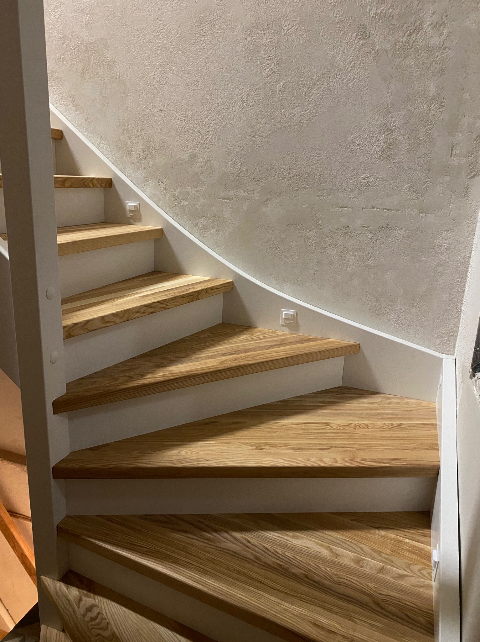 Spiralne schody z drewna o jasnym odcieniu, z białymi podstopnicami i oświetleniem LED w ścianie, prowadzące ku górze wzdłuż teksturowanej ściany.