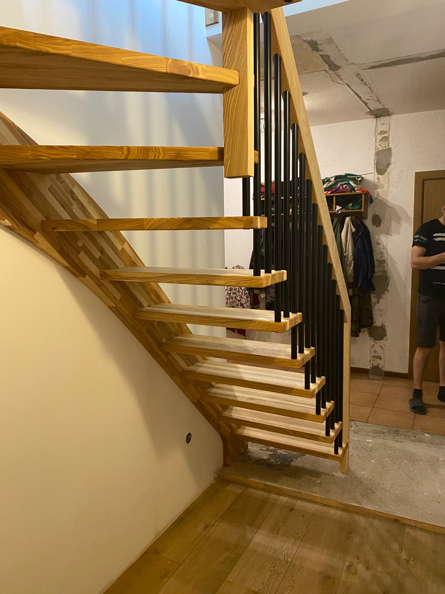 Jasne, drewniane schody z czarną, metalową balustradą, widoczne z boku. W tle fragment pomieszczenia w trakcie wykańczania, z widoczną instalacją na suficie.