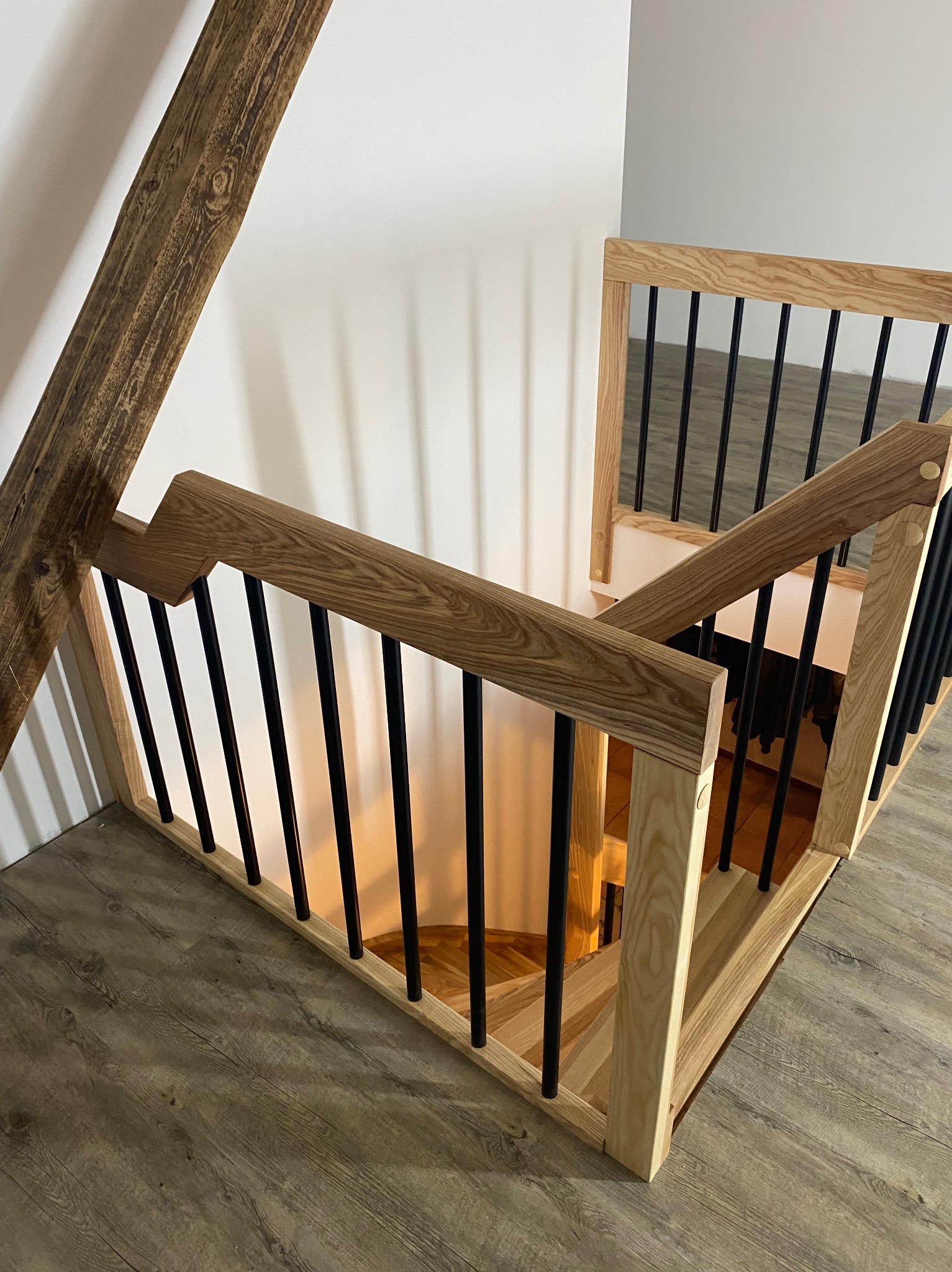 Spiralne schody z jasnego drewna z czarnymi, metalowymi prętami balustrady, widoczne z góry, z fragmentem belki stropowej w lewym górnym rogu.