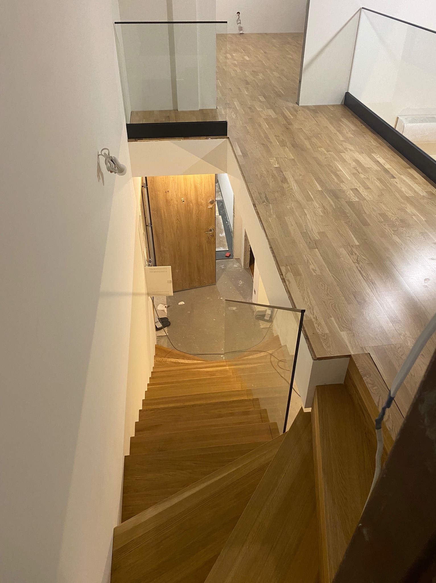 Wnętrze domu z drewnianymi schodami i szklaną balustradą. Widok z góry na dół, ukazujący drewniane stopnie i podłogę, oraz fragment drzwi wejściowych.