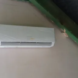 Biała jednostka klimatyzacji wewnętrznej, owinięta folią ochronną, zamontowana na kremowej ścianie pod skośnym, zielonym elementem konstrukcyjnym dachu, obok okna.