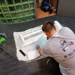 Mężczyzna w szarej koszulce z logo firmy MONKIPC montuje filtr do rozłożonej jednostki klimatyzacyjnej na podłodze, obok leży zielony odkurzacz i narzędzia.