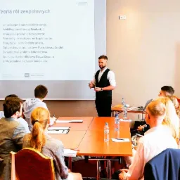 Prelegent w czarnej kamizelce gestykuluje podczas prezentacji o teoriach ról zespołowych przed grupą słuchaczy siedzących przy stole z butelkami wody.