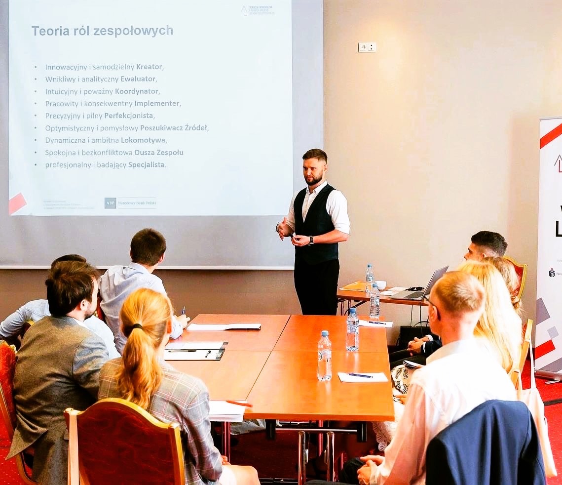 Prelegent w czarnej kamizelce gestykuluje podczas prezentacji o teoriach ról zespołowych przed grupą słuchaczy siedzących przy stole z butelkami wody.