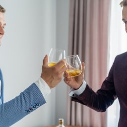 FFWStudio - Dwóch elegancko ubranych mężczyzn wznosi toast szklankami z bursztynowym napojem z lodem, jeden w błękitnej marynarce w drobną kratę z ozdobnymi guzikami, drugi w bordowym smokingu z czarną muchą.