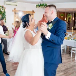 Uśmiechnięta para młoda w tańcu, on w granatowym garniturze z butonierką, ona w białej sukni z welonem, w tle goście weselni i dekoracje sali.