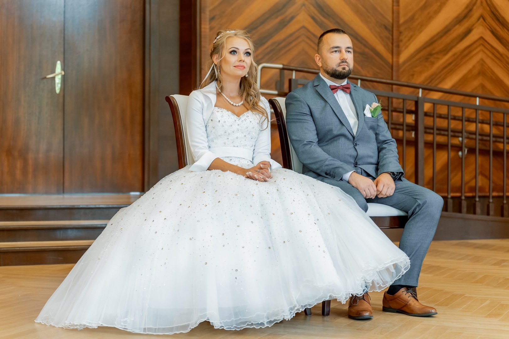 Para młoda siedzi na krzesłach podczas ceremonii, panna młoda w sukni z błyszczącymi aplikacjami i bolerku, pan młody w szarym garniturze z bordową muchą, w tle drewniane panele i schody.