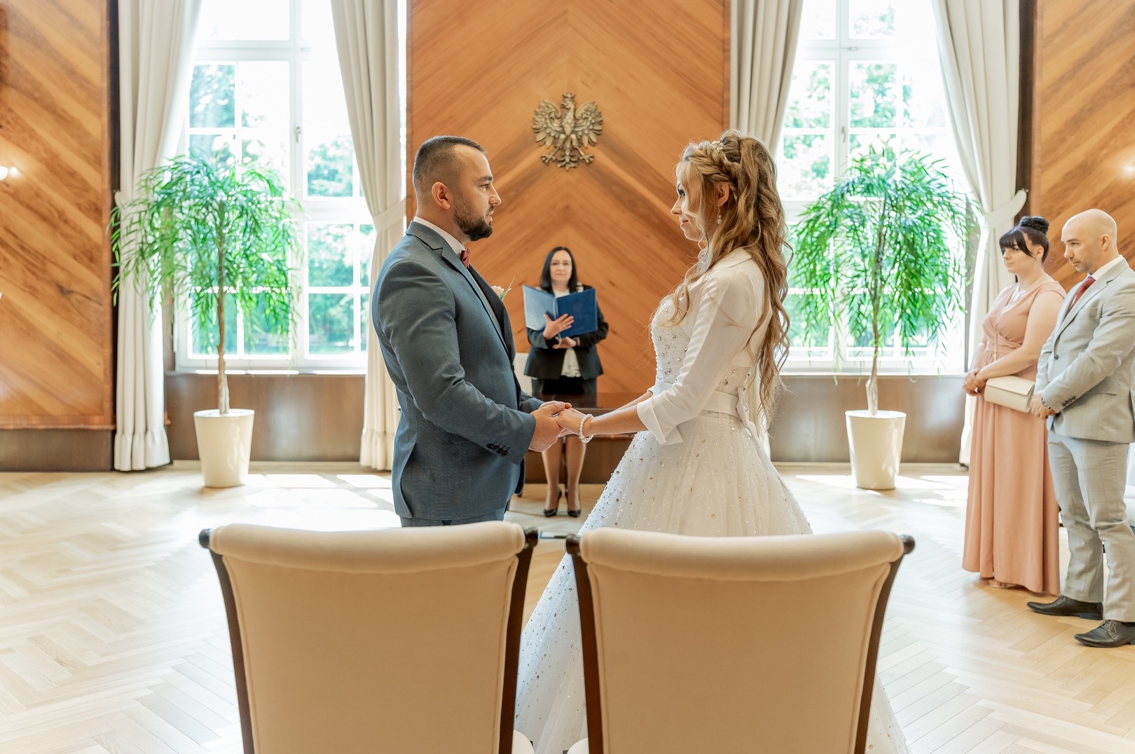 Para młoda trzymająca się za ręce podczas ceremonii ślubnej w jasnym wnętrzu, na tle świadków i urzędniczki, z dekoracyjnymi roślinami w donicach i godłem państwowym na ścianie.