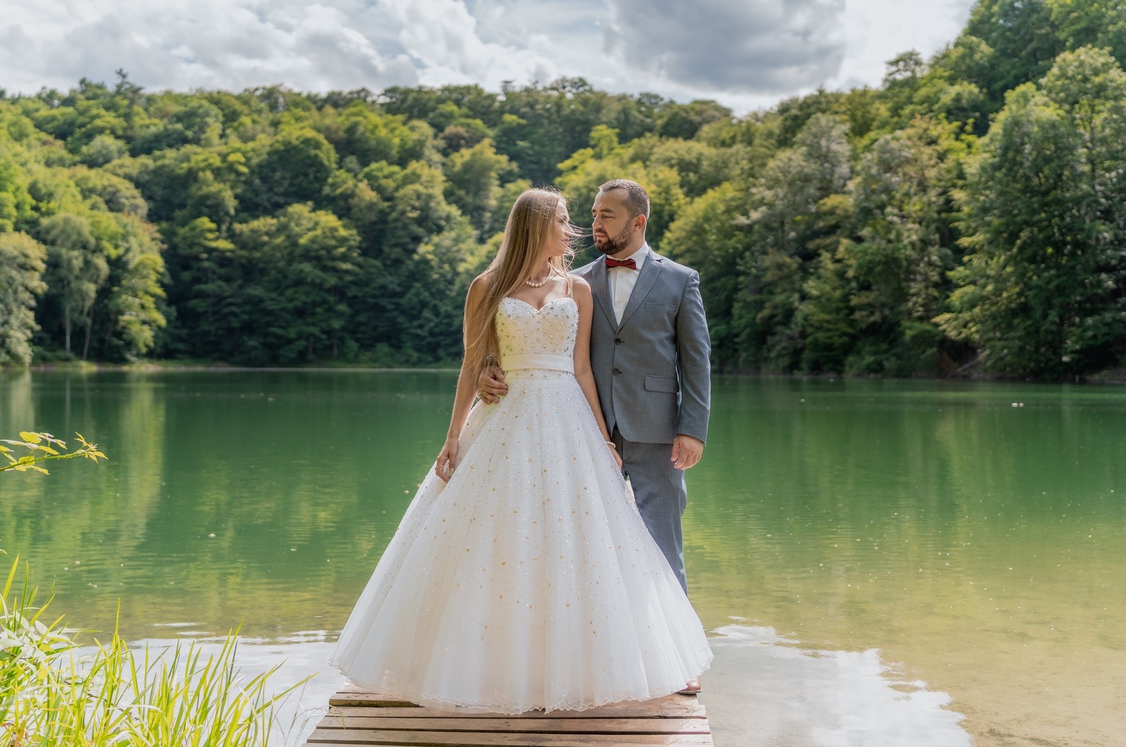 Para młoda, panna młoda w sukni z kryształkami i pan młody w szarym garniturze z muchą, pozują na drewnianym pomoście nad zielonym jeziorem, otoczeni lasem.
