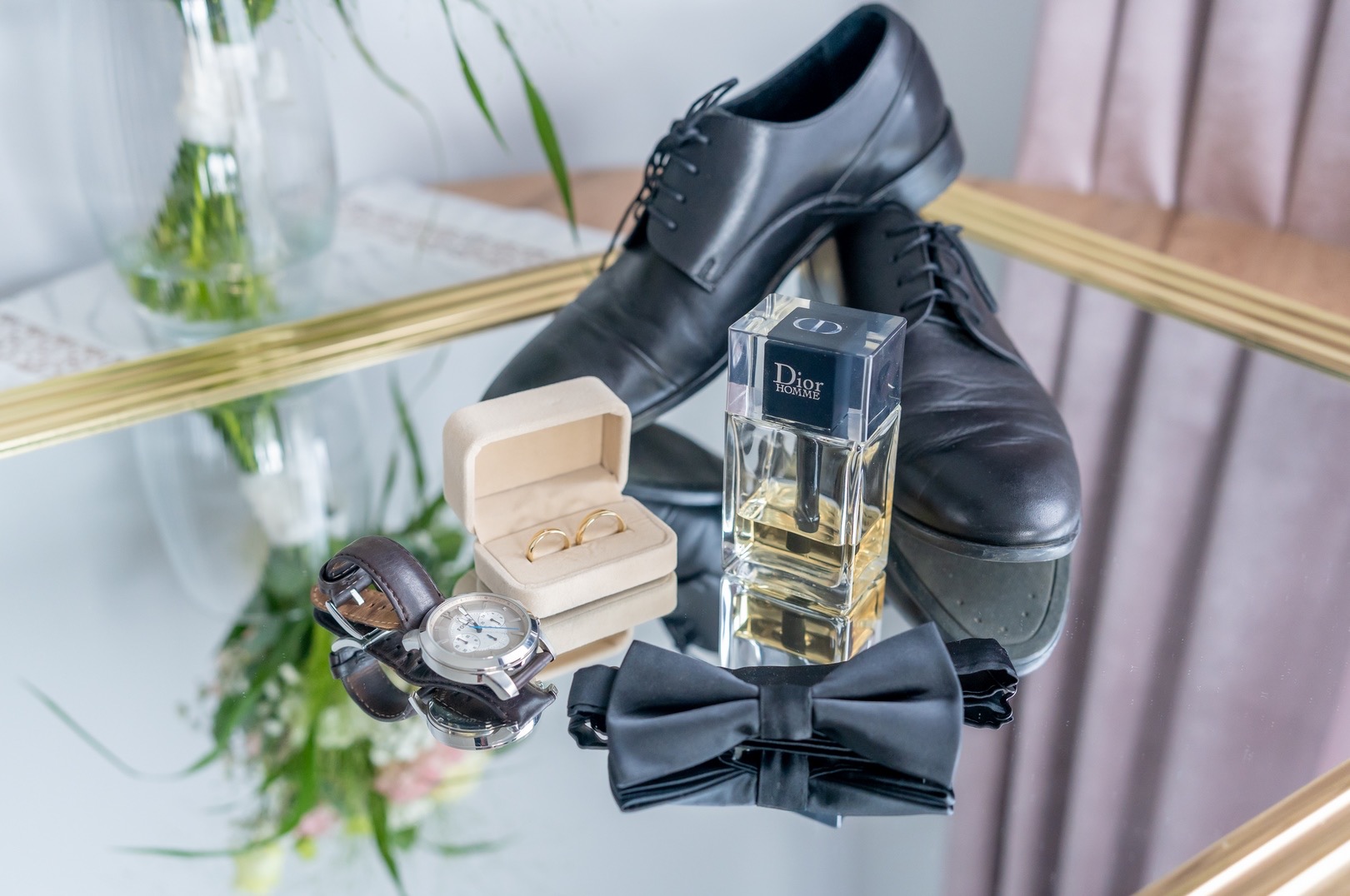 Męskie akcesoria ślubne ułożone na lustrzanej powierzchni: czarne skórzane buty, muszka, zegarek z brązowym paskiem, flakon perfum Dior Homme i pudełko z obrączkami.