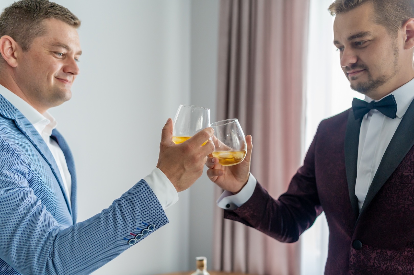 Dwóch elegancko ubranych mężczyzn wznosi toast szklankami z bursztynowym napojem z lodem, jeden w błękitnej marynarce w drobną kratę z ozdobnymi guzikami, drugi w bordowym smokingu z czarną muchą.