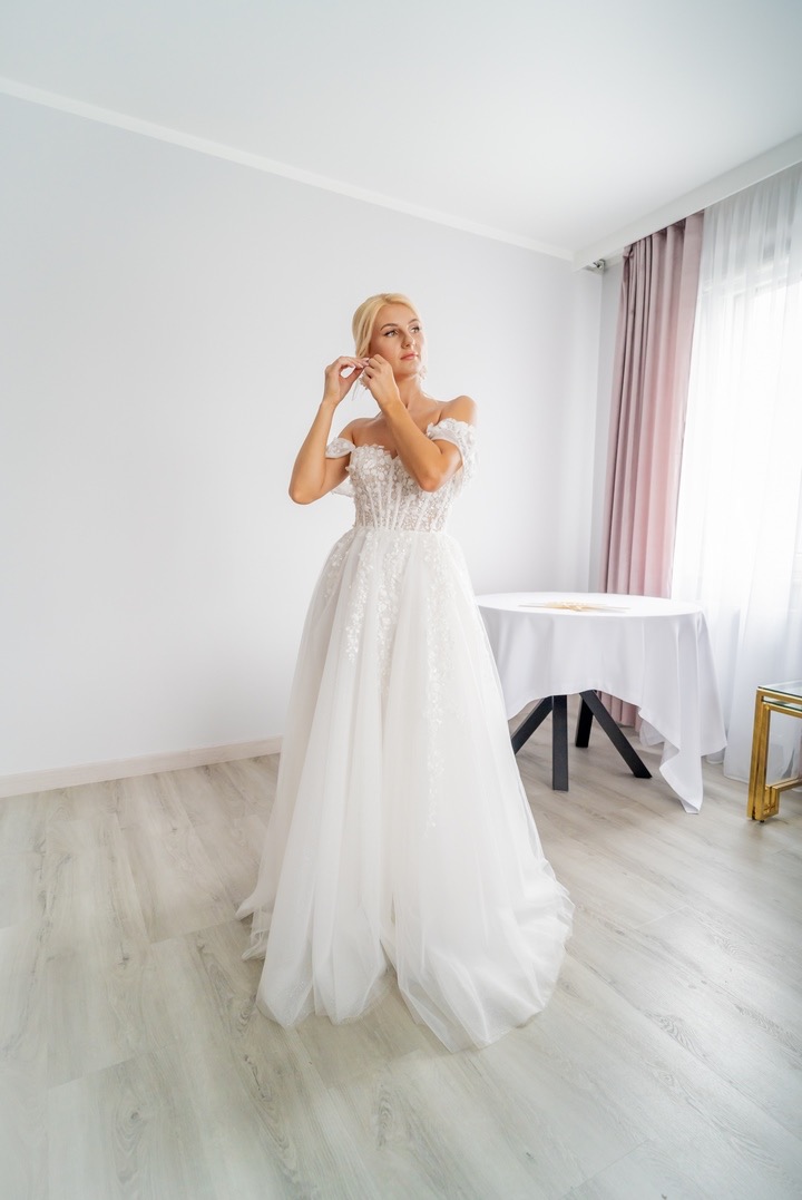 Panna młoda w białej sukni z odkrytymi ramionami, dopinająca kolczyk przed ślubem, w tle stół z białym obrusem i zasłony w odcieniach różu.