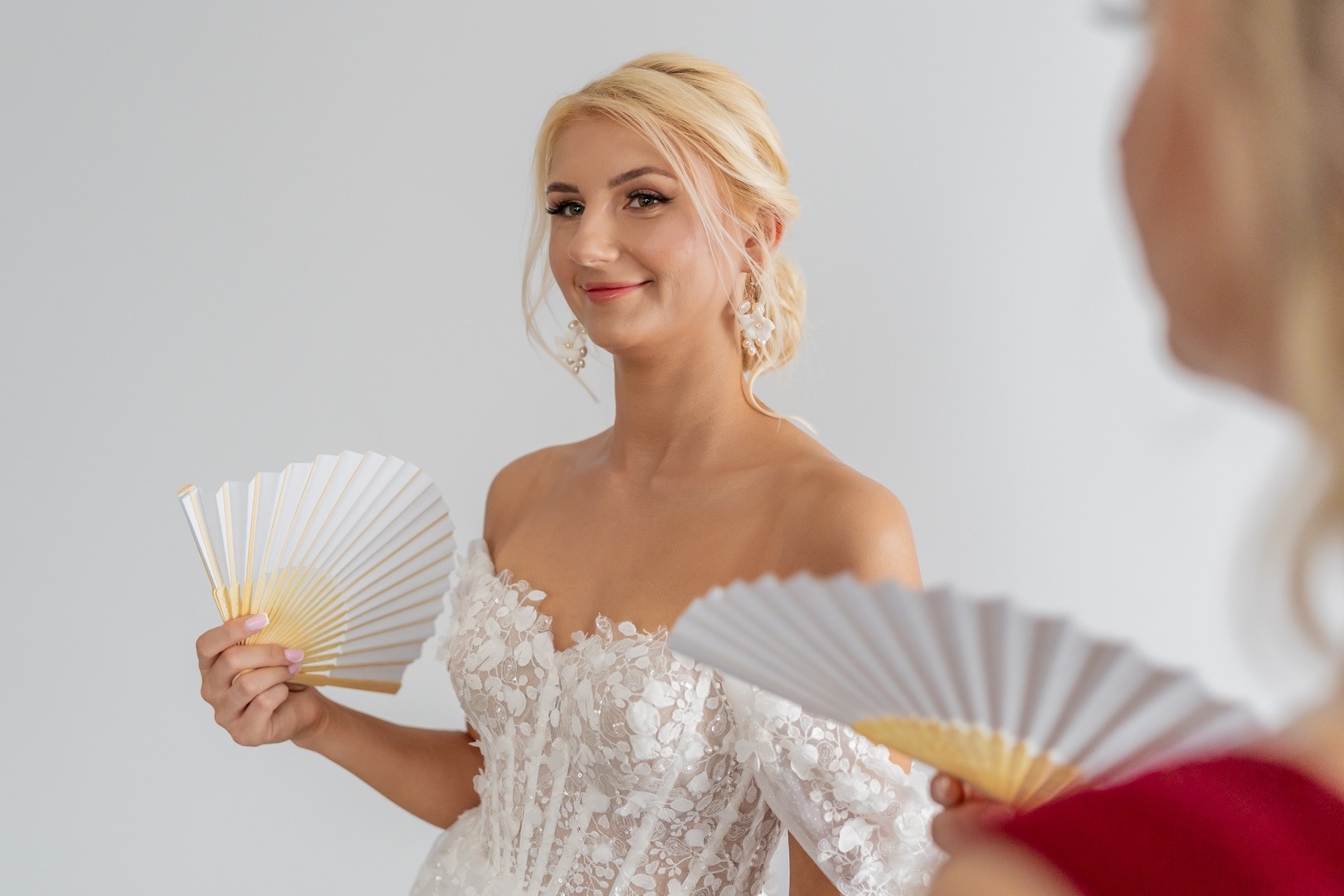 Panna młoda w koronkowej sukni trzyma biały wachlarz, odbicie w lustrze z drugim wachlarzem, kolczyki z motywem kwiatowym, upięte blond włosy.