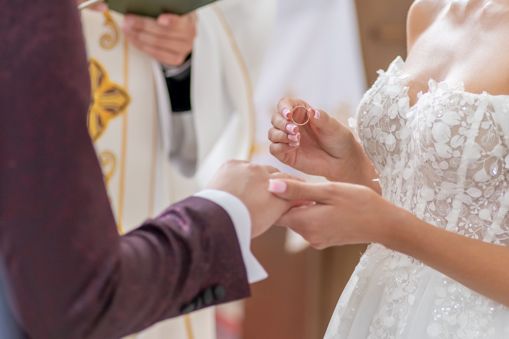 Szczecin: Detal podczas ceremonii ślubnej, panna młoda w koronkowej sukni trzyma obrączkę nad dłonią pana młodego w bordowym garniturze, w tle ksiądz w białej szacie.