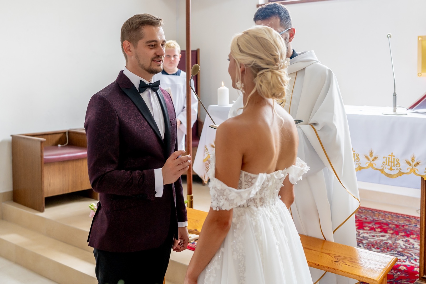 Para młoda podczas ceremonii ślubnej w kościele: pan młody w bordowym smokingu z czarną muchą, panna młoda w białej sukni z odkrytymi ramionami, w tle ksiądz i ministrant.