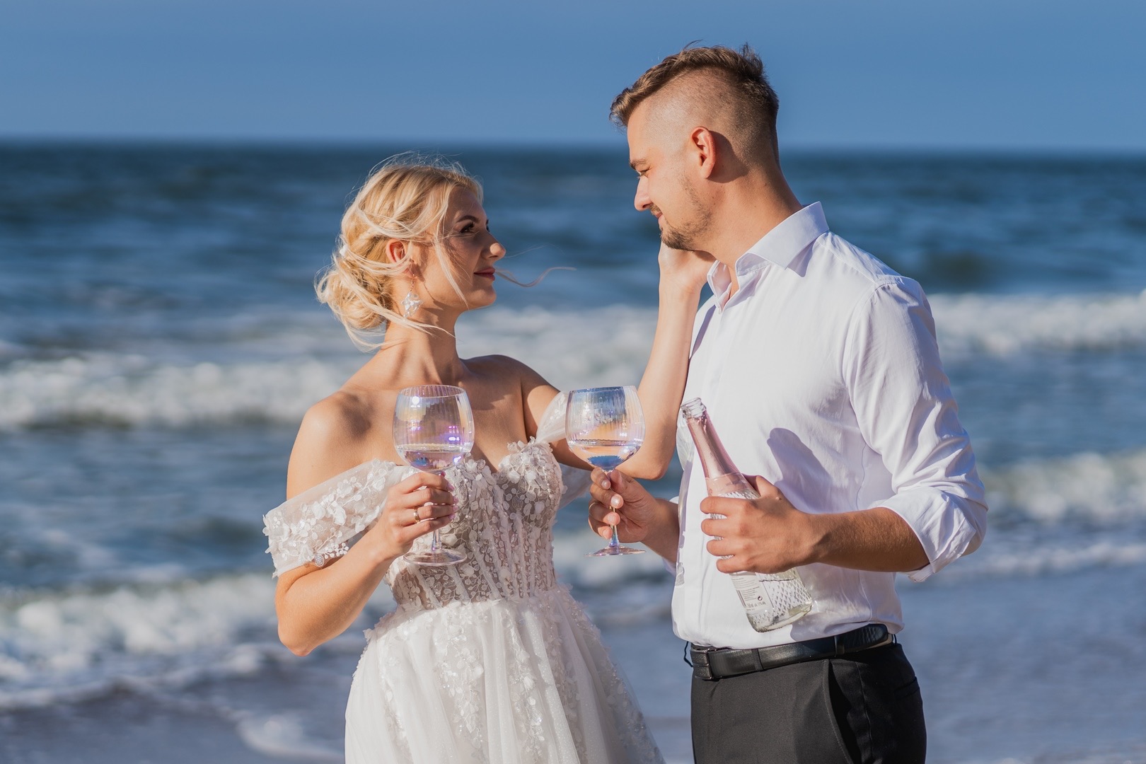 Para młoda w strojach ślubnych na plaży w słoneczny dzień, trzyma kieliszki z napojem i butelkę szampana, patrząc na siebie z uśmiechem.