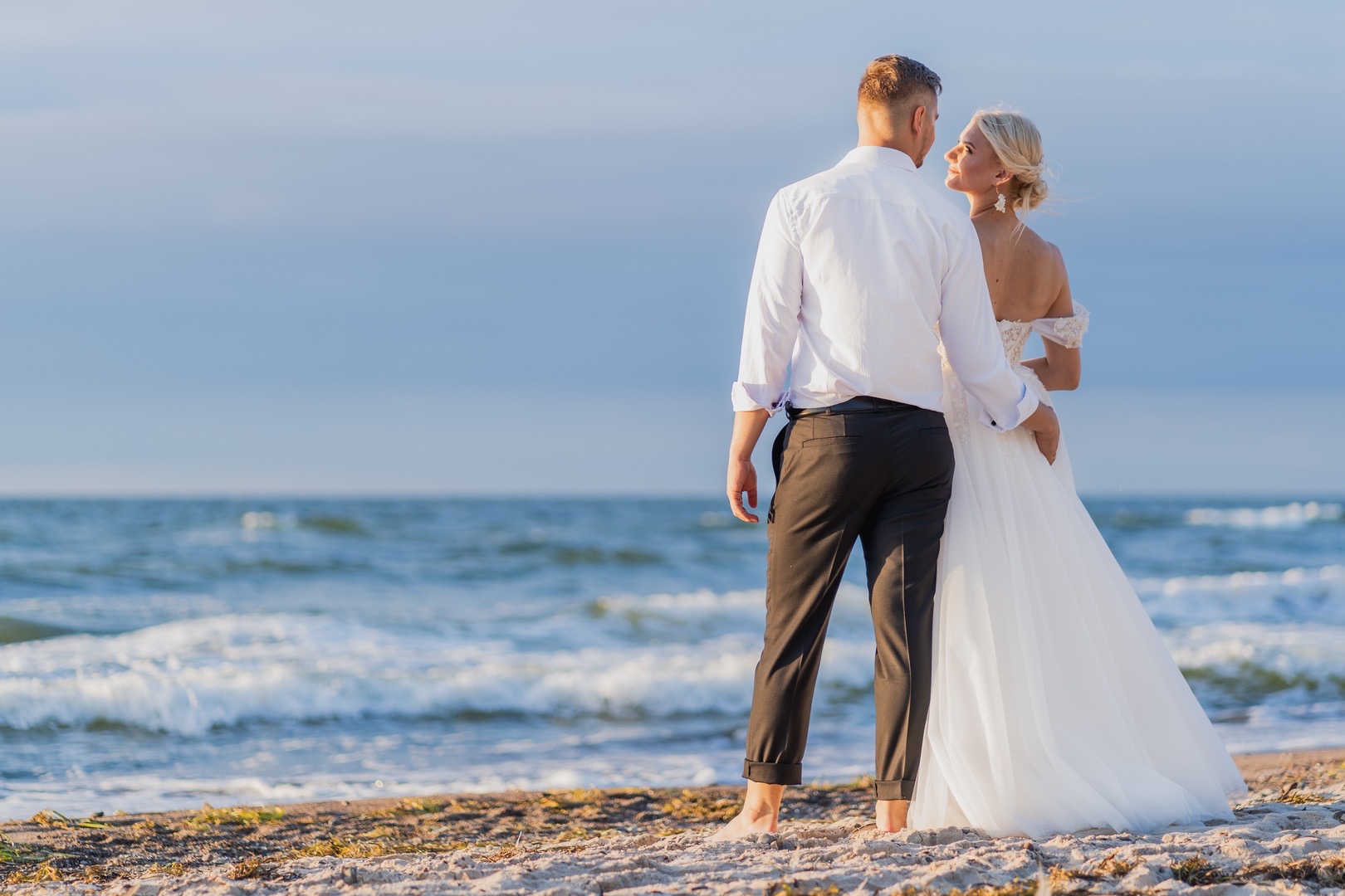 Para młoda, on w białej koszuli i ciemnych spodniach, ona w białej sukni ślubnej z odkrytymi ramionami, stoją na plaży tyłem do widza, patrząc na siebie na tle morza i nieba.