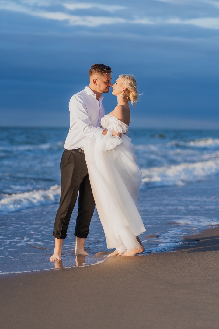 Para młoda na plaży podczas zachodu słońca; panna młoda w białej sukni z koronkową górą i upiętymi blond włosami, pan młody w białej koszuli i czarnych spodniach, oboje boso stoją w wodzie, prawie...