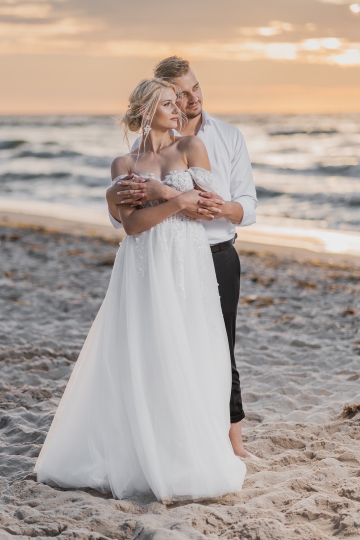 Para młoda na plaży o zachodzie słońca: panna młoda w sukni z odkrytymi ramionami i delikatnymi zdobieniami, pan młody w białej koszuli obejmuje ją od tyłu, oboje patrzą w dal w kierunku morza.