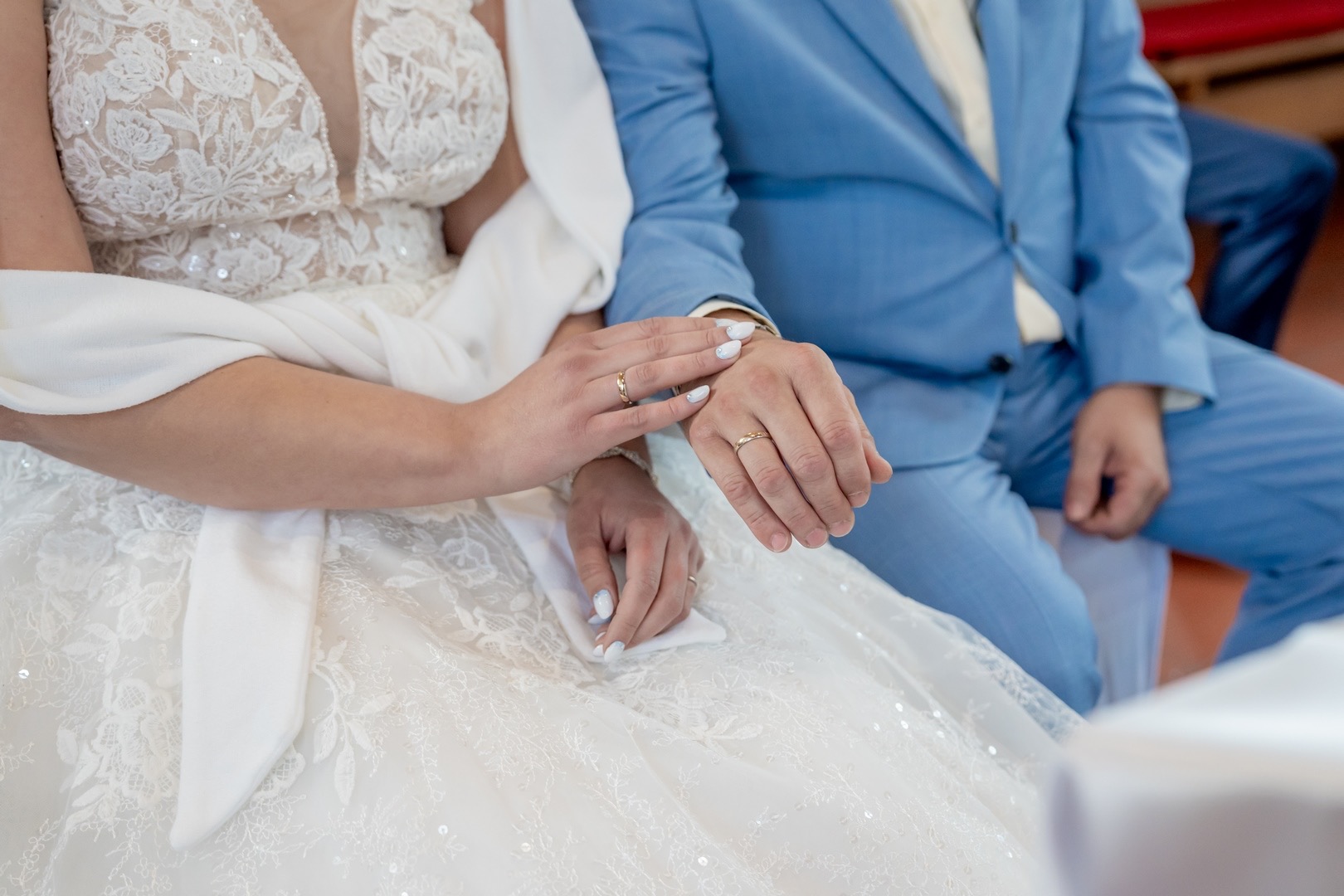 Dłonie nowożeńców z obrączkami na tle koronkowej sukni ślubnej i błękitnego garnituru.