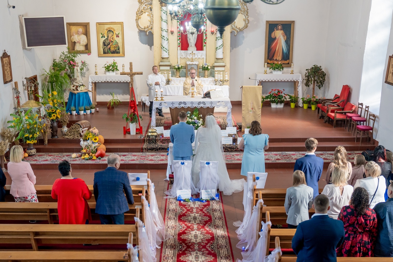 Para młoda podczas ceremonii ślubnej w kościele, widok zza pleców na ołtarz, księdza i gości, dekoracje kwiatowe i jesienne akcenty.