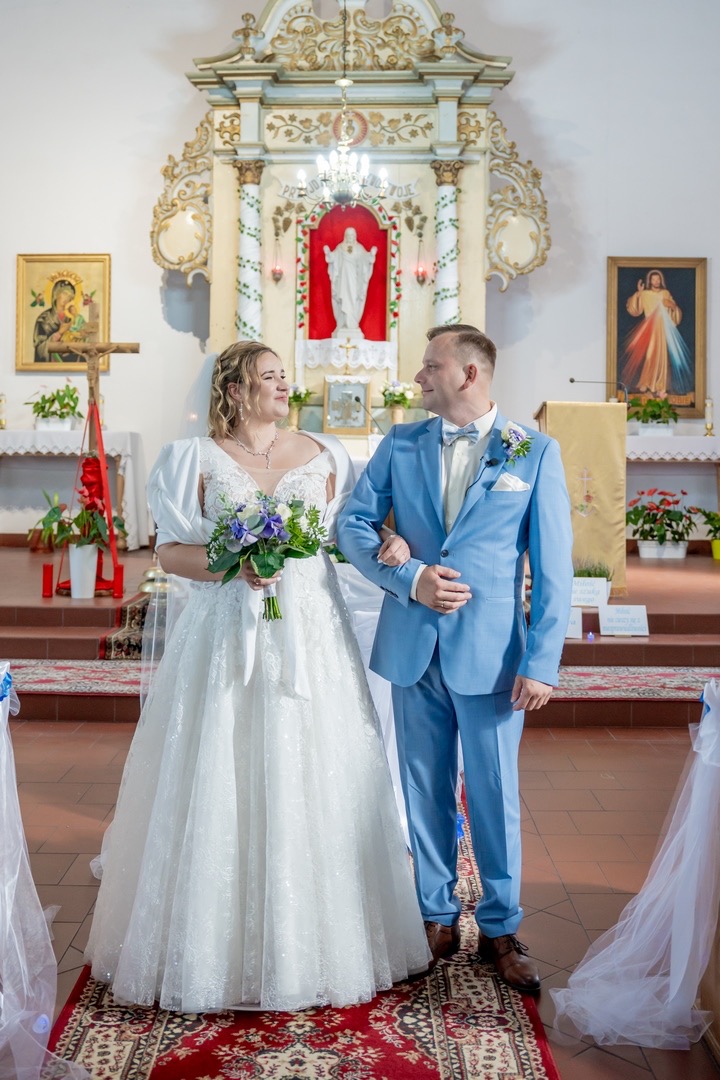 Para młoda, panna młoda w białej sukni z bukietem i pan młody w błękitnym garniturze, stoją na czerwonym dywanie w kościele, patrząc na siebie z uśmiechem, na tle ołtarza z figurą.