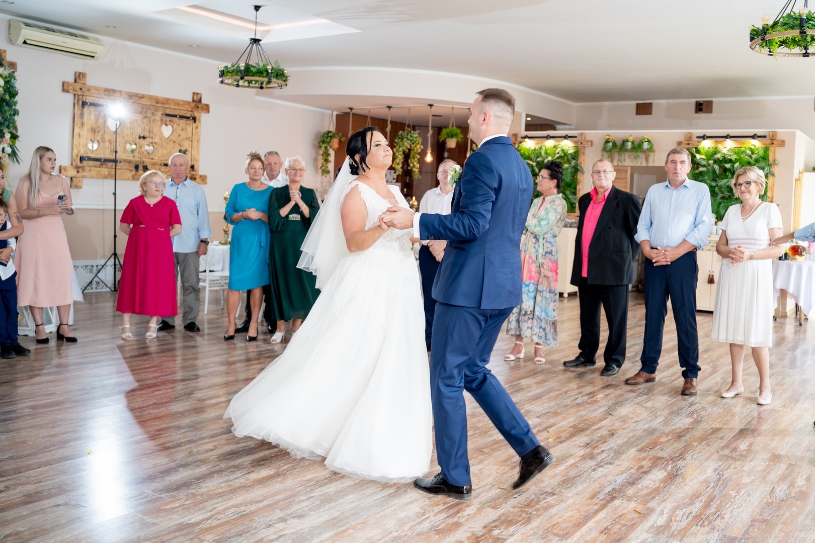 Para młoda w pierwszym tańcu na parkiecie, otoczona gośćmi weselnymi w jasnym, przestronnym wnętrzu sali bankietowej z drewnianymi dekoracjami i roślinami.