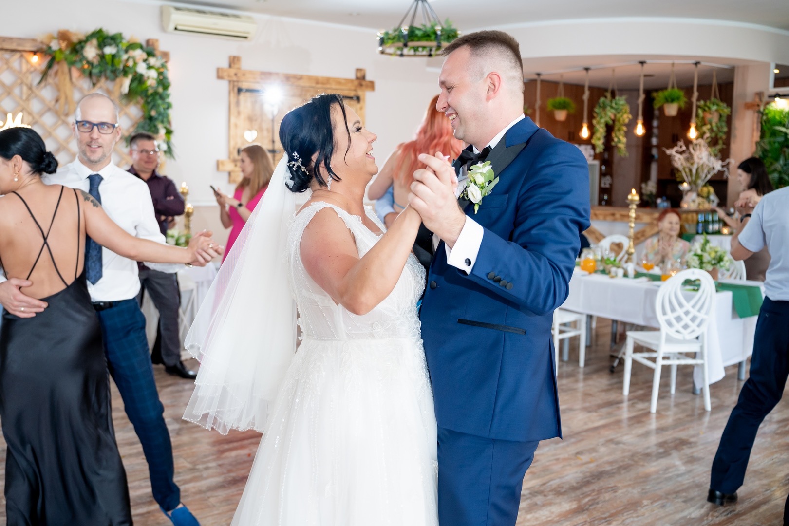 Uśmiechnięta para młoda w tańcu, on w granatowym garniturze z butonierką, ona w białej sukni z welonem, w tle goście weselni i dekoracje sali.